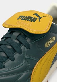 PUMA KING-sneakeren har en grønn lær-overdel, gule detaljer på tungen, teksturerte lisser og gyldne merkevaredetaljer.