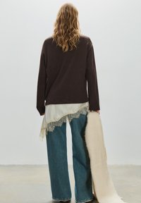 Barna hosszú ujjú felső, krémszínű csipkés szegélyű blúz fölött, széles szárú kék farmernadrággal, kezében egy krémszínű, texturált darabot tartva.