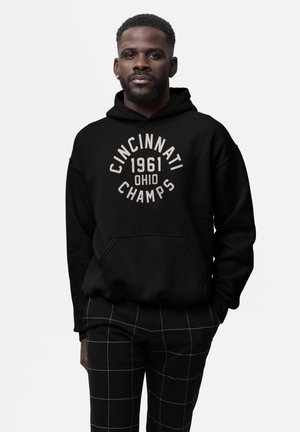 Uomo con capelli corti, barba e orecchino che indossa una felpa nera con il testo "Cincinnati 1961 Ohio Champs" e pantaloni neri con un motivo a griglia bianca.