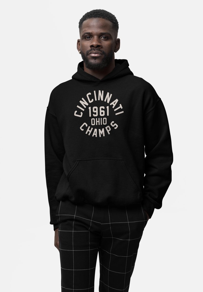 Uomo con capelli corti, barba e orecchino che indossa una felpa nera con il testo "Cincinnati 1961 Ohio Champs" e pantaloni neri con un motivo a griglia bianca.