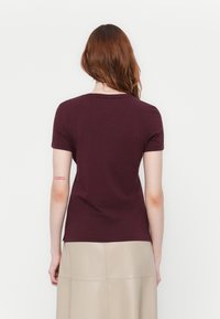 Mujer con largo cabello rojo viste una blusa de manga corta color burdeos y una falda beige, de pie de espaldas contra un fondo blanco liso.