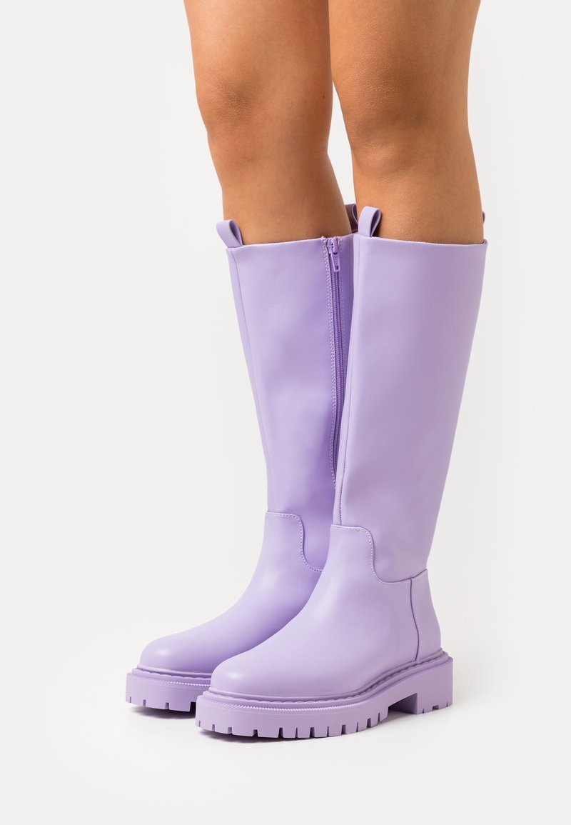 Bottes en caoutchouc lavande montant jusqu'aux genoux, à texture lisse, fermeture éclair latérale et semelle épaisse avec motif de crampons. Elles comportent des languettes de traction en haut.