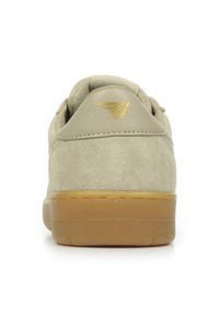 Zapatilla de ante beige con un dorso liso, acento del logo dorado y suela de goma. Diseñada para uso casual, con forma redondeada y detalles texturizados.