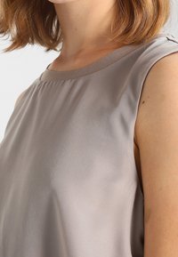Ärmelloses Oberteil aus weichem, hellem Taupe-Stoff mit glatter Textur und geripptem Ausschnitt, der subtile Nähdetails aufweist.
