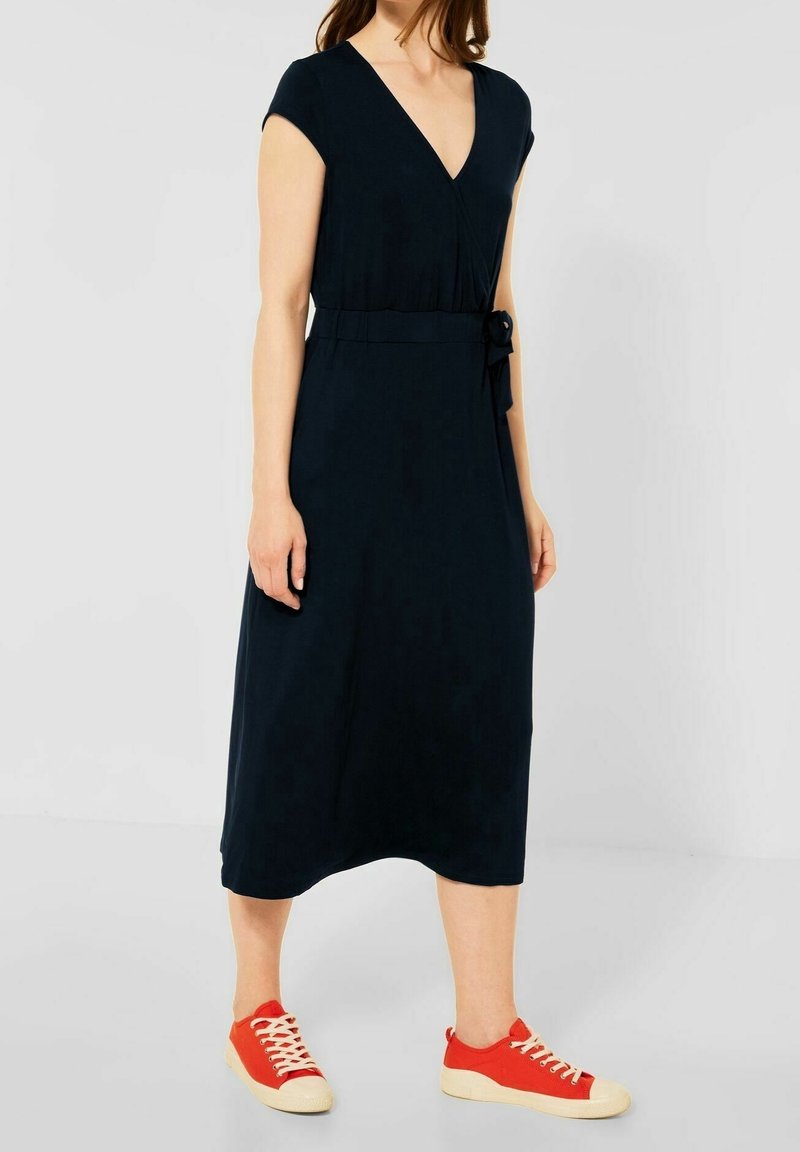 Granatowa sukienka do kolan z głębokim dekoltem w serek, krótkimi rękawami i wiązaniem w talii. Połączona z czerwonymi trampkami i kremowymi podeszwami.