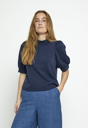 Kobieta o długich blond włosach nosi granatową bluzkę z bufkami i wysokiej talii teksturowane, szerokie niebieskie spodnie, stoi z jedną ręką w kieszeni.
