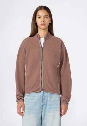 Junge Frau trägt eine braune Fleecejacke mit Reißverschluss und hellblaue Jeans und steht vor einem einfarbigen hellen Hintergrund.