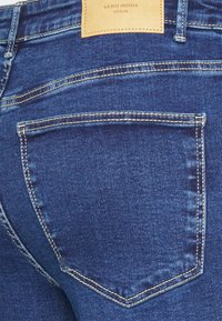 Vaqueros de mezclilla azul oscuro con costuras beige en contraste, una etiqueta de cuero en la cinturilla y un detalle de un solo bolsillo trasero.