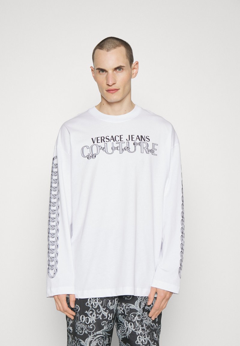Camiseta blanca de manga larga con el logo "VERSACE JEANS COUTURE" en negro, que presenta un patrón gráfico de cadenas a lo largo de las mangas y un corte relajado.