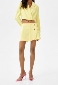 Cropped yellow blazer with wide lapels, paired with a short yellow skirt featuring two gold buttons and a wrap design. Pink strappy heels.