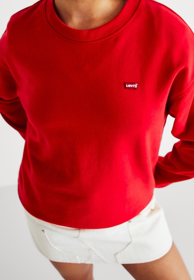 Personne portant un sweat-shirt Levi's rouge et une jupe blanche, debout avec les mains derrière le dos sur un fond uni.