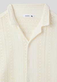 Camicetta bianca a maniche corte realizzata in tessuto a maglia testurizzata, caratterizzata da un colletto classico, chiusura a bottoni e motivi intricati su tutta la superficie.