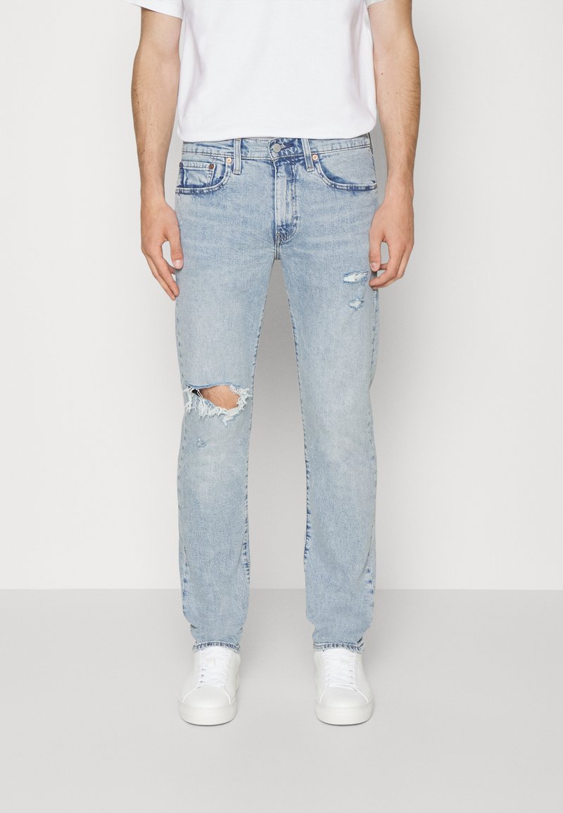 Lichtblauwe denim jeans met een rechte snit, voorzien van versleten details, waaronder een gat op de knie. Gecombineerd met een eenvoudig wit t-shirt en witte sneakers.