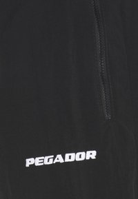 Czarna kurtka z widocznym zamkiem błyskawicznym oraz haftowanym logo "PEGADOR" w białym kolorze. Materiał wydaje się gładki i lekki.
