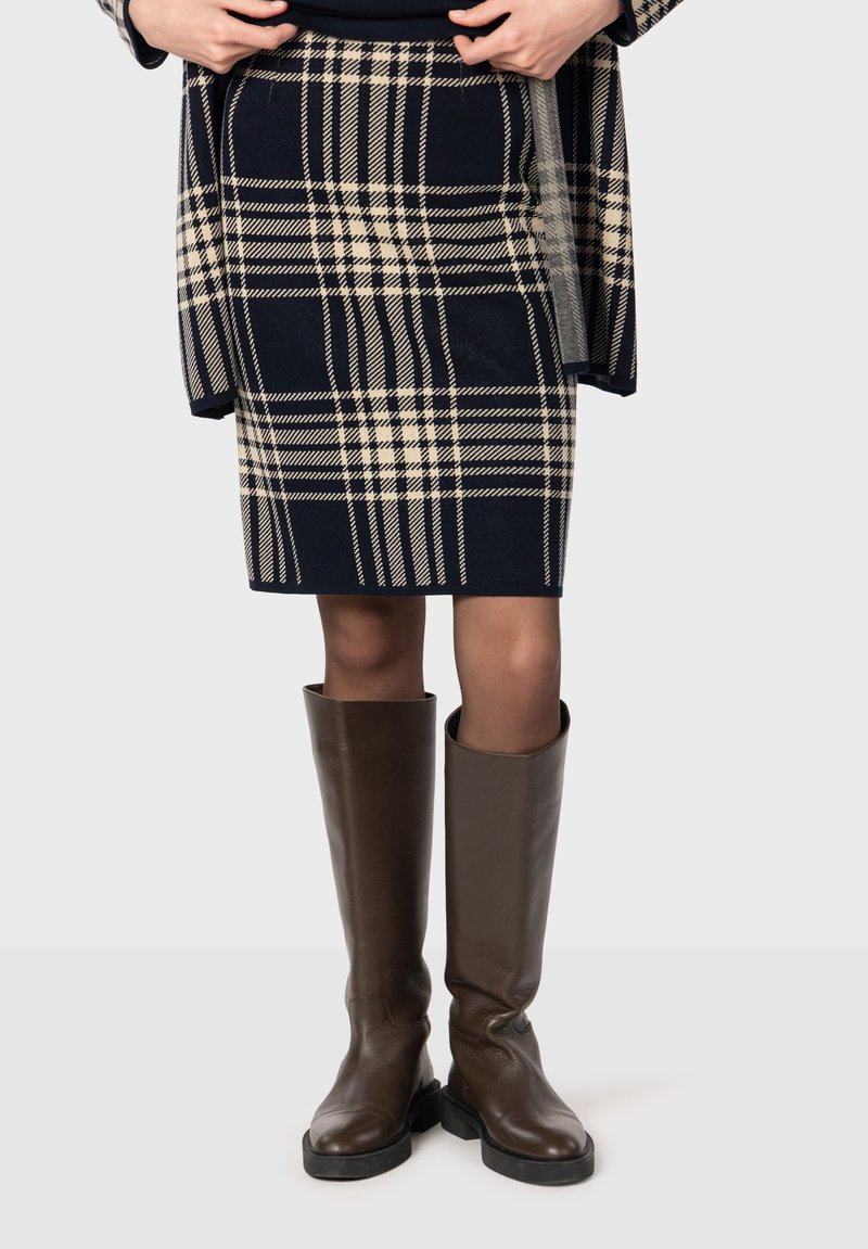 Granatowa spódnica w kratę z kremowymi akcentami, do kolan, uszyta z teksturowanej tkaniny. Połączona z brązowymi skórzanymi butami do kolan z czarną podeszwą.