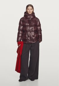 Bordeauxrode donzen jas met een hoge kraag, glanzende textuur en zakken aan de voorkant; model houdt een rode sjaal vast; gecombineerd met losse zwarte broek.