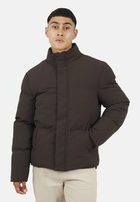 Giacca a vento marrone scuro con colletto alto, texture a coste, zip frontale e tasche laterali. Indossata sopra pantaloni chiari.
