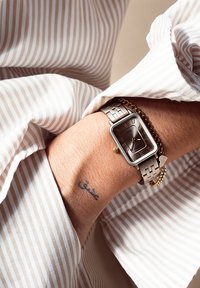Montre rectangulaire en argent avec un cadran marron texturé et des chiffres romains, ornée d'un bracelet en chaîne dorée. Portée au poignet.