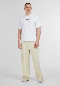 Tricou alb din bumbac, cu mâneci scurte și un logo negru "KANI". Asortat cu pantaloni lejeri crem și adidași bej.