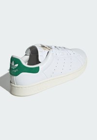 Białe skórzane sneakersy z zielonym zamszowym zakładką na pięcie, perforowanymi detalami i beżową gumową podeszwą. Posiadają złote logo na języku.