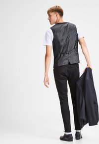 Chaleco de satén negro con una sola correa en la espalda, llevado sobre una camisa blanca, combinado con pantalones negros y sosteniendo una chaqueta oscura, visto desde detrás.