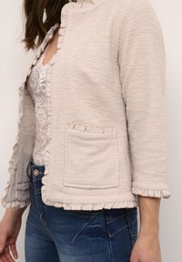 Veste texturée beige clair avec des bords froncés, poche ornée et manches 3/4, associée à un haut en dentelle blanche et un jean bleu.