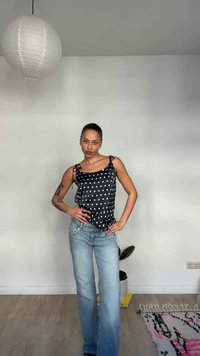 Femme posant à l'intérieur portant un haut sans manches noir à pois et un jean bleu clair, avec un luminaire et un tapis visibles dans la pièce.