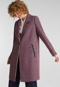 Kvinde med krøllet hår iført en lang rød, hvid og marineblå houndstooth-mønstret frakke over en hvid skjorte og mørke bukser, stående med armene strakt ud.