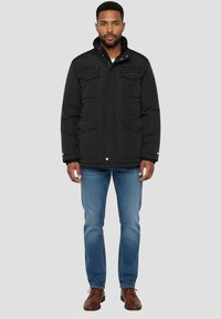 Giacca nera isolata con zip frontale, due tasche sul petto e bottoni a scatto; indossata con jeans blu e scarpe marroni. Texture liscia.