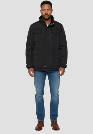 Zwarte geïsoleerde jas met een ritssluiting aan de voorkant, twee borstzakken en drukknoopsluitingen; gecombineerd met een blauwe spijkerbroek en bruine schoenen. Glad oppervlak.