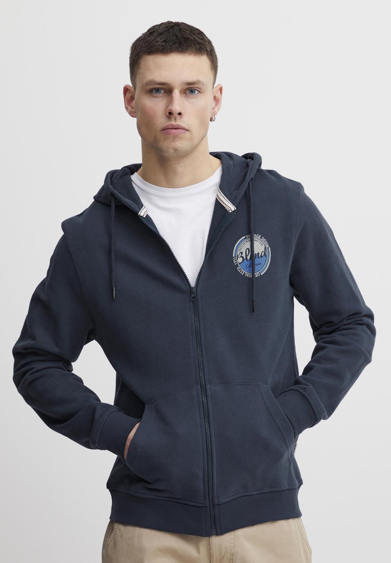 Mornarska zip-up dukserica s prednjim logotipom, kapuljačom sa šniranjem i kengurskim džepovima. Sadrži rebraste manžete i donji rub. Mekani materijal od pamučne mješavine.