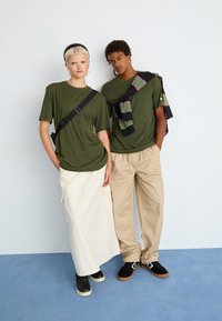 Dwójka młodych dorosłych w oliwkowo-zielonych koszulach i beżowych spodniach stoi przy białej ścianie na niebieskiej podłodze.