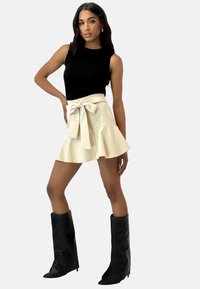Top negro de canalé sin mangas, falda corta acampanada de color crema con lazo en la cintura, combinada con botas negras hasta la rodilla con punteras puntiagudas.