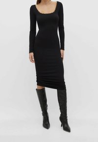 Schwarzes langärmliges Kleid mit quadratischem Ausschnitt, der mit Spitze verziert ist, figurbetontem Schnitt und Raffung am Saum. Getragen mit knielangen schwarzen Stiefeln.