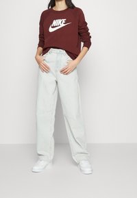 Personne portant un sweat à capuche Nike bordeaux, un jean ample bleu clair et des baskets blanches, se tenant contre un mur blanc.