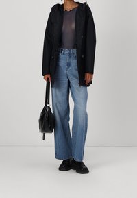 Černá voděodolná bunda s kapucí, průsvitný černý top, světle modré široké kalhoty s vysokým pasem, černá taška a robustní černé boty.