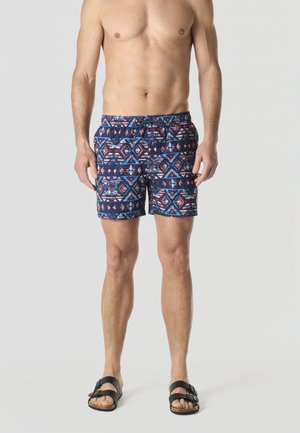 Hombre con pantalones cortos de baño azules con estampado y sandalias negras, de pie contra un fondo liso y claro.