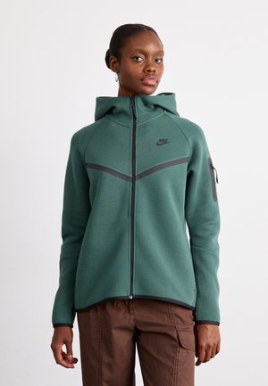 Felpa con zip Nike di colore verde scuro in pile con dettagli neri, caratterizzata da un colletto alto, zip frontale e tasche laterali. Texture liscia.