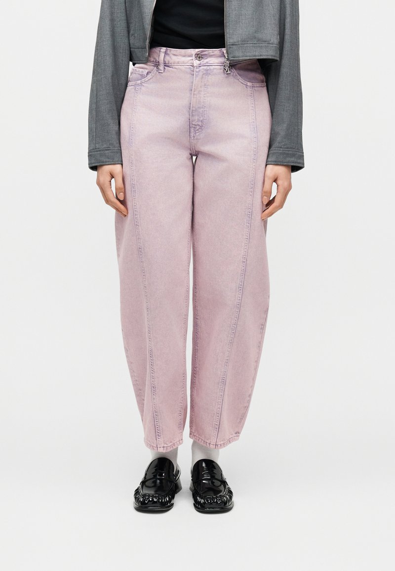 Γυναίκα που φοράει ανοιχτό ροζ τζιν παντελόνι ψηλής μέσης με ραφές μπροστά, γκρι κοντό σακάκι, λευκές κάλτσες και μαύρα γυαλιστερά μοκασίνια, στέκεται μπροστά σε λευκό φόντο.