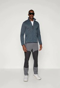 Muž v tmavých slnečných okuliaroch, modrom fleecovom zipsovom saku, šedých a čiernych nohaviciach a bielych teniskách stojaci na pozadí so svetlým povrchom.