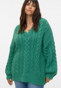 Pull en tricot vert surdimensionné avec un col en V, présentant des motifs texturés et des poignets côtelés, porté sur un jean bleu.