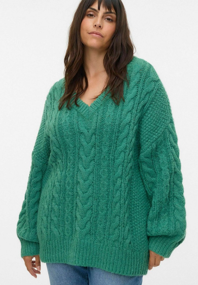 Pull en tricot vert surdimensionné avec un col en V, présentant des motifs texturés et des poignets côtelés, porté sur un jean bleu.