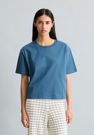 Kobieta z długimi ciemnymi włosami nosi niebieską koszulkę z krótkim rękawem i białe spodnie z niebieskim geometrycznym wzorem, stoi na tle jasnoszarego tła.