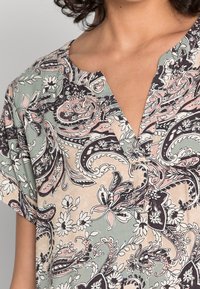 Mujer vistiendo una blusa de manga corta con un patrón de paisley y flores en verde, rosa y beige, y un cuello en V con abertura.