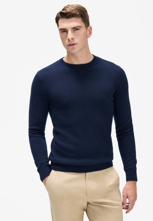 Marinblå crew-neck sweater, tætsiddende design, blød tekstur, lange ærmer, ribbet kant og manchetter, parret med beige bukser.