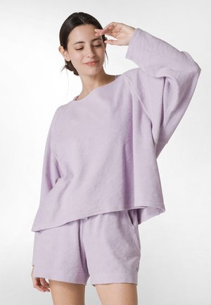 Completo felpa di colore lilla chiaro, in tessuto morbido, composto da una maglietta a maniche lunghe dal taglio ampio e pantaloni corti a vestibilità rilassata con tasche laterali e orli leggermente sfilacciati.