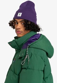 Chaqueta tipo puffer verde con acentos morados en los hombros, que presenta un alto cuello, botones a presión y un cordón ajustable. Gorro morado con etiqueta de logo.