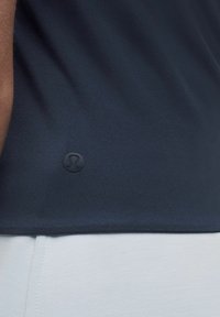 Marinblått atletiskt linne med slät yta och en figurnära design, med en liten svart logotyp. Den nedre fållen överlappar vit textil.