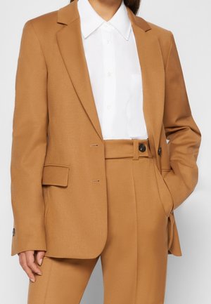 Woman wearing a camel brown tailored suit with blazer and high-waisted pants over a white collared button-up shirt.