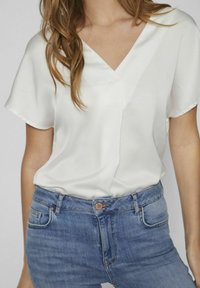 Blusa bianca a maniche corte e scollo a V realizzata in un tessuto liscio, abbinata a jeans di denim blu chiaro con vita standard e dettagli delle tasche.